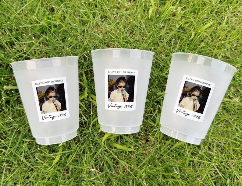 May include: Three frosted plastic cups with labels featuring a photo and text. The labels read "Riley's 30th Birthday" and "Vintage 1995." The cups are arranged on a bed of green grass.