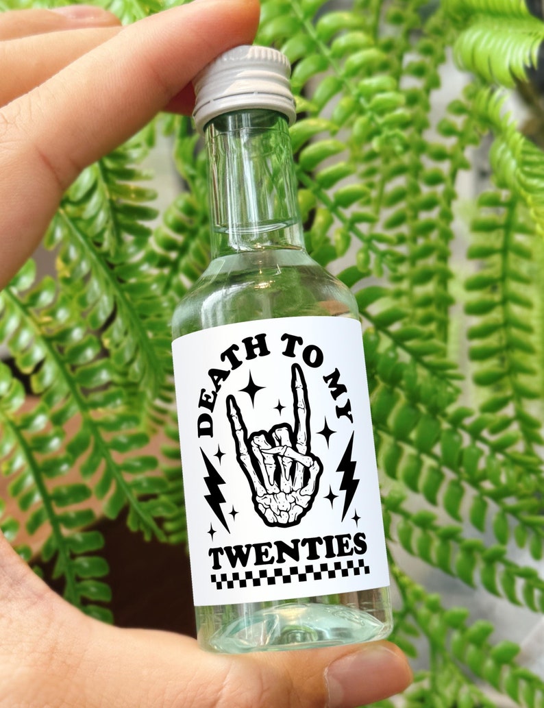 May include: A small clear glass bottle with a white label that says "Death to My Twenties". The label has a black and white illustration of a skeleton hand making a rock and roll hand gesture.