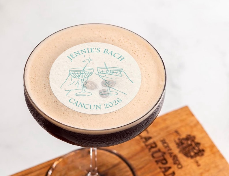 May include: A close-up of a cocktail with a white sticker on top. The sticker has a drawing of two martini glasses and the text "Jennie's Bach Cancun 2026".