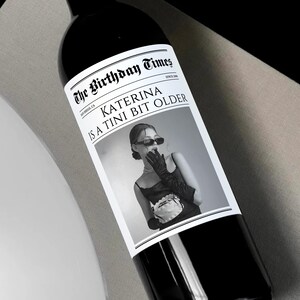 May include: A black and white photo of a woman with a surprised expression on her face. The photo is on a wine bottle label with the text "The Birthday Times" and "Katerina is a Tini Bit Older".