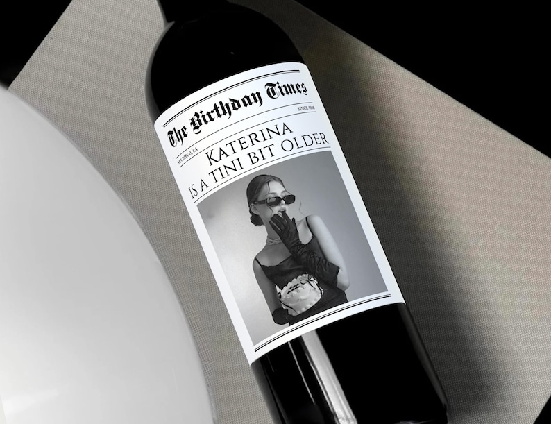 May include: A black and white photo of a woman with a surprised expression on her face. The photo is on a wine bottle label with the text "The Birthday Times" and "Katerina is a Tini Bit Older".