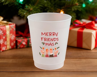 May include: A white plastic cup with the words "MERRY FRIENDS MAS" in red script. The cup has a festive design with illustrations of people toasting with champagne glasses. Christmas-themed gift boxes and a decorated tree are in the background.