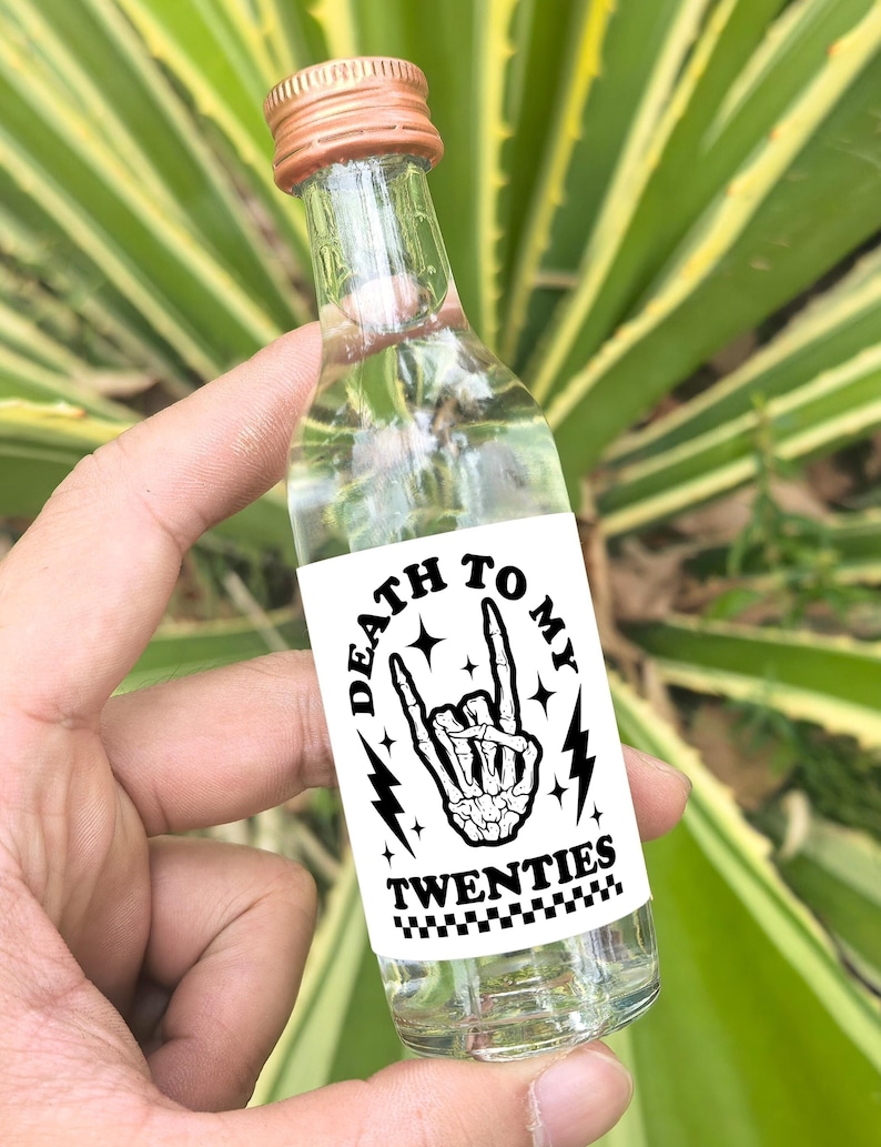 May include: A clear glass bottle with a copper-colored cap and a white label that says "Death to My Twenties" with a black and white illustration of a skeleton hand making a rock and roll hand gesture.
