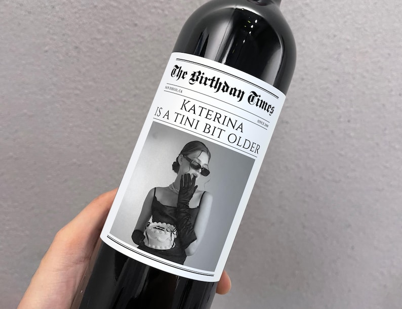 May include: A black and white photo of a woman wearing sunglasses and a black dress holding a cake. The photo is on a wine bottle label with the text "The Birthday Times" and "Katerina is a Tini Bit Older".