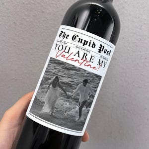 May include: A dark red wine bottle with a custom label. The label reads "The Cupid Post" with the headline "YOU ARE MY Valentine!" in red script. A black and white photo of a couple in the ocean is below the headline.