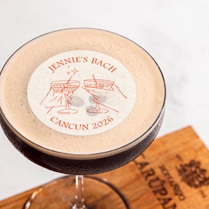May include: A close-up of a cocktail with a white sticker on top. The sticker has a red design with two hands holding martini glasses and the text "JENNIE'S BACH CANCUN 2026".