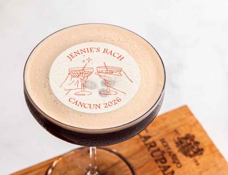 May include: A close-up of a cocktail with a white sticker on top. The sticker has a red design with two hands holding martini glasses and the text "JENNIE'S BACH CANCUN 2026".