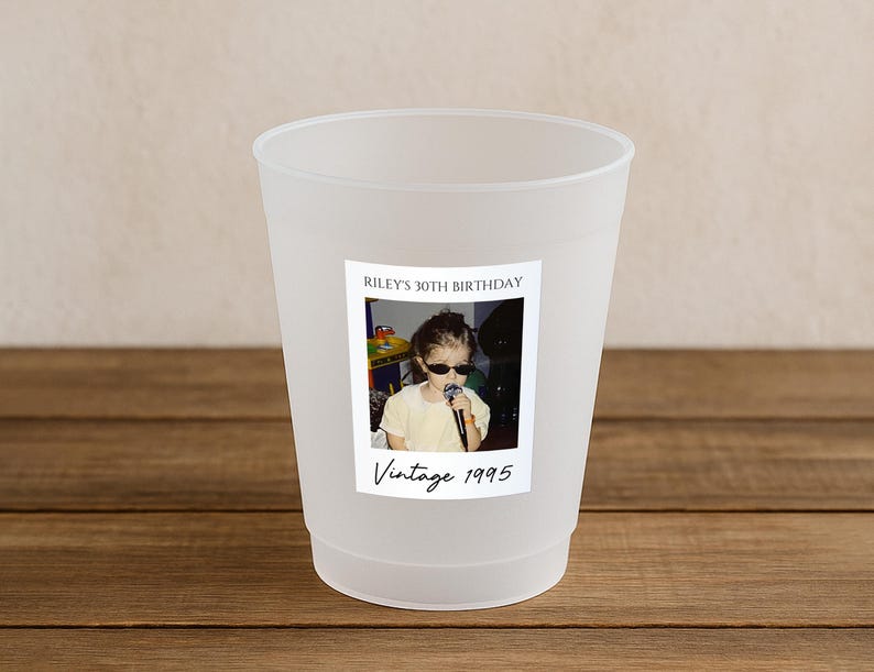 May include: A frosted plastic cup with a photo label. The label reads "Riley's 30th Birthday" and "Vintage 1995" with a photo of a child wearing sunglasses and holding a microphone. The cup is on a wooden surface.