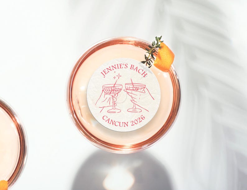 May include: A white round sticker with pink text that says "Jennie's Bach Cancun 2026" and a pink illustration of two hands holding up martini glasses. The sticker is on top of a glass of pink liquid.