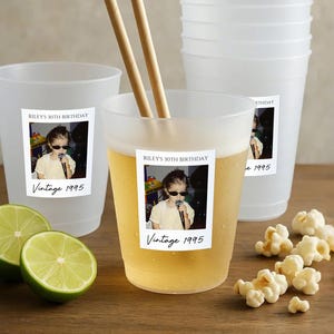 May include: Frosted plastic cups with a "Riley's 30th Birthday Vintage 1995" sticker. One cup contains a light-colored beverage and wooden chopsticks. Limes and popcorn are on a wooden surface.
