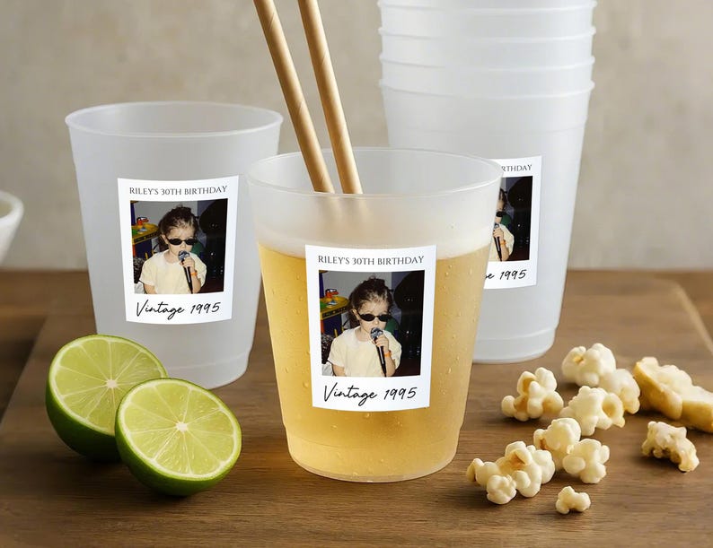 May include: Frosted plastic cups with a "Riley's 30th Birthday Vintage 1995" sticker. One cup contains a light-colored beverage and wooden chopsticks. Limes and popcorn are on a wooden surface.