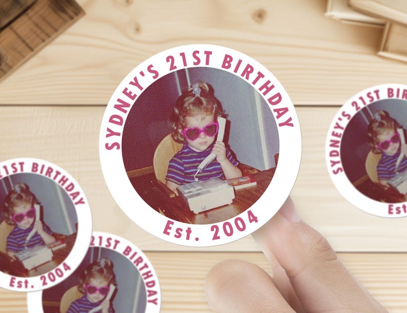 Op de afbeelding: Een witte ronde sticker met een roze cirkel in het midden. De roze cirkel heeft de tekst "Sydney's 21st Birthday" in roze letters. Binnen de roze cirkel is een foto van een jong meisje met roze zonnebril, aan de telefoon. De tekst "Est. 2004" staat onder de roze cirkel.