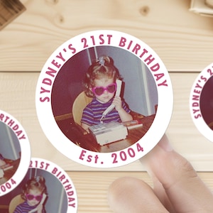 Op de afbeelding: Een witte ronde sticker met een roze cirkel in het midden. De roze cirkel heeft de tekst "Sydney's 21st Birthday" in roze letters. Binnen de roze cirkel is een foto van een jong meisje met roze zonnebril, aan de telefoon. De tekst "Est. 2004" staat onder de roze cirkel.