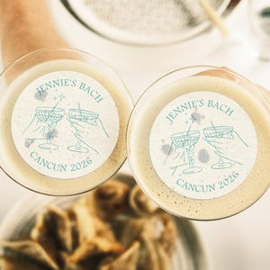 May include: Two white cocktail coasters with a blue design of two martini glasses clinking together. The text on the coasters reads "Jennie's Bach Cancun 2026".