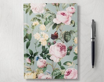 May include: A floral journal with a pen. The journal features a light blue background with a pattern of pink roses, yellow flowers, green leaves, and butterflies. A silver and black pen rests beside the journal.