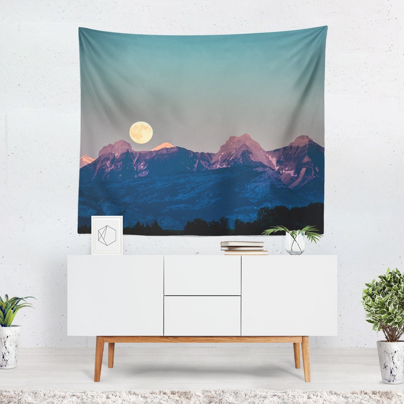 May include: A wall tapestry featuring a mountain range under a full moon. The mountains are a deep blue with pink highlights, and the moon is a bright yellow. The sky transitions from a light blue to a darker gray. A white cabinet with wooden legs is below the tapestry.