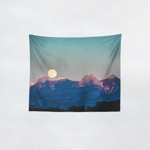 May include: A wall tapestry featuring a scenic mountain landscape under a full moon. The mountains are a deep blue with touches of pink on the peaks, set against a gradient sky of teal and gray. The moon is a bright yellow circle.