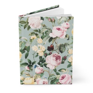 May include: A hardcover journal with a floral design featuring pink and yellow roses, green leaves, and butterflies on a light blue background. The journal is open, revealing blank white pages.