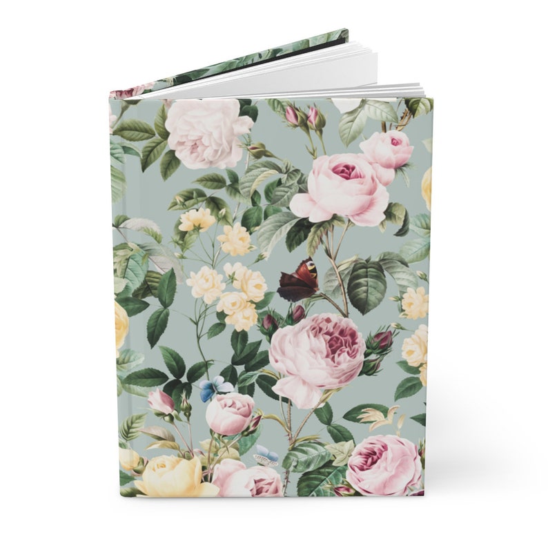 May include: A hardcover journal with a floral design featuring pink and yellow roses, green leaves, and butterflies on a light blue background. The journal is open, revealing blank white pages.