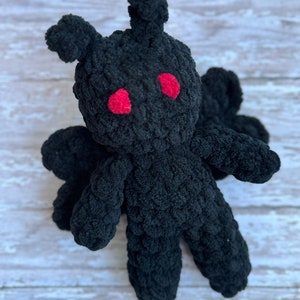 May include: Black crocheted plush toy with red eyes. The toy has a humanoid shape with long arms and legs.