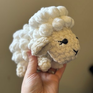 May include: A white crocheted sheep plush toy with black eyes. The sheep has a fluffy white wool texture and a beige face.