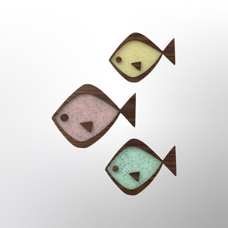 Puede incluir: Tres decoraciones de peces de madera con un dise&ntilde;o moderno de mediados de siglo. Cada pez tiene un marco de madera marr&oacute;n oscuro y un interior moteado en colores pastel: amarillo, rosa y azul. Los peces tienen una forma simple y geom&eacute;trica con cola y un ojo peque&ntilde;o.