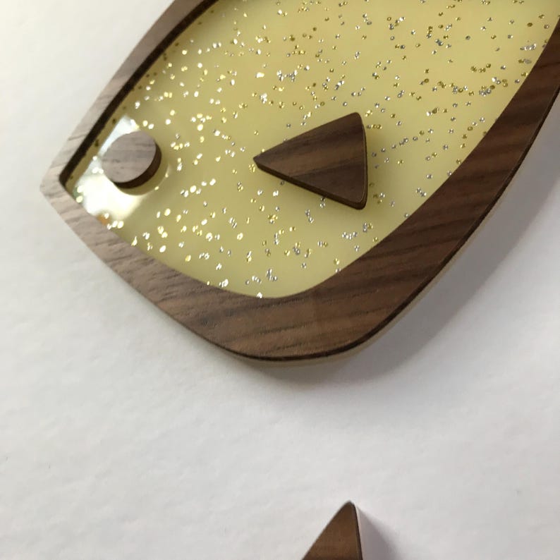 Puede incluir: Una pieza decorativa de arte mural con forma de pez de madera. La pieza presenta un fondo amarillo claro con purpurina plateada, un marco de madera oscura y detalles de madera. El dise&ntilde;o incluye un elemento de madera circular y triangular.