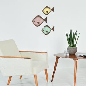Puede incluir: Tres adornos de pared de peces de madera en colores pastel: amarillo, rosa y turquesa. Cada pez tiene un contorno y una aleta caudal de color marr&oacute;n oscuro. Los peces est&aacute;n montados en una pared blanca, encima de un sill&oacute;n blanco y una mesa auxiliar de madera.