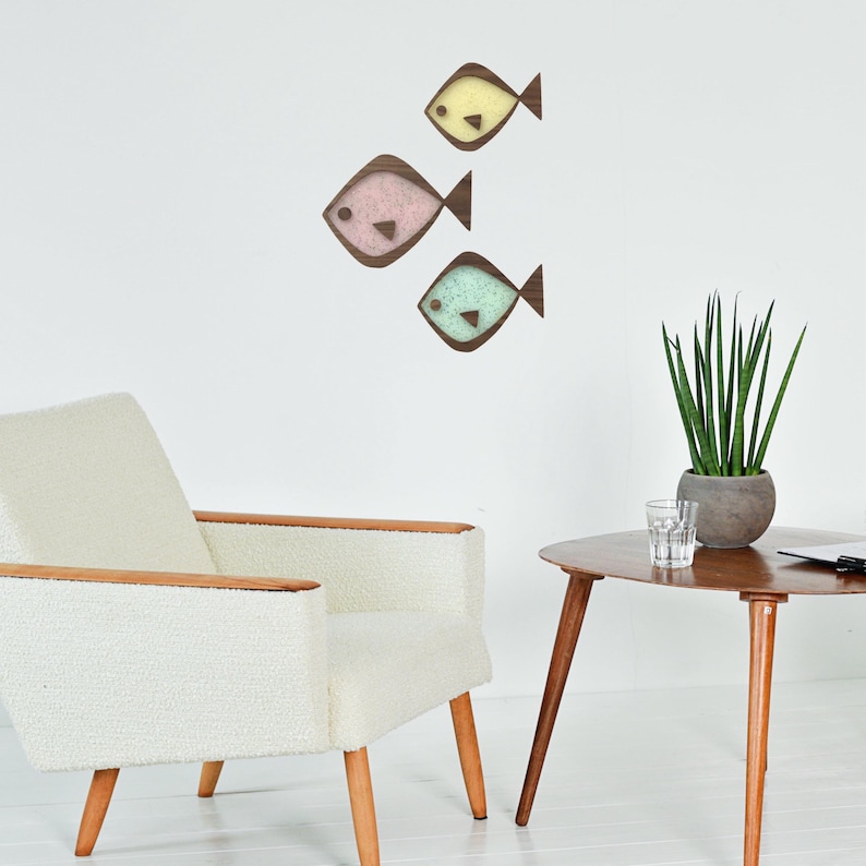 Puede incluir: Tres adornos de pared de peces de madera en colores pastel: amarillo, rosa y turquesa. Cada pez tiene un contorno y una aleta caudal de color marr&oacute;n oscuro. Los peces est&aacute;n montados en una pared blanca, encima de un sill&oacute;n blanco y una mesa auxiliar de madera.