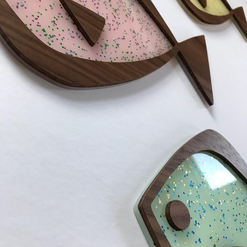 Puede incluir: Arte mural con forma de pez de madera con un dise&ntilde;o moderno de mediados de siglo. Los peces tienen un marco de madera marr&oacute;n oscuro y un centro de acr&iacute;lico transparente lleno de purpurina de colores. El fondo es blanco.