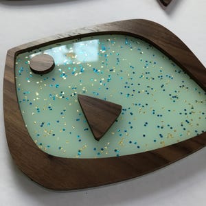 Puede incluir: Una bandeja decorativa de madera con un dise&ntilde;o &uacute;nico en forma de ojo. La bandeja presenta un interior verde claro con detalles de purpurina azul y dorada, enmarcado por un borde de madera oscura. Incluye un peque&ntilde;o c&iacute;rculo y tri&aacute;ngulo de madera.