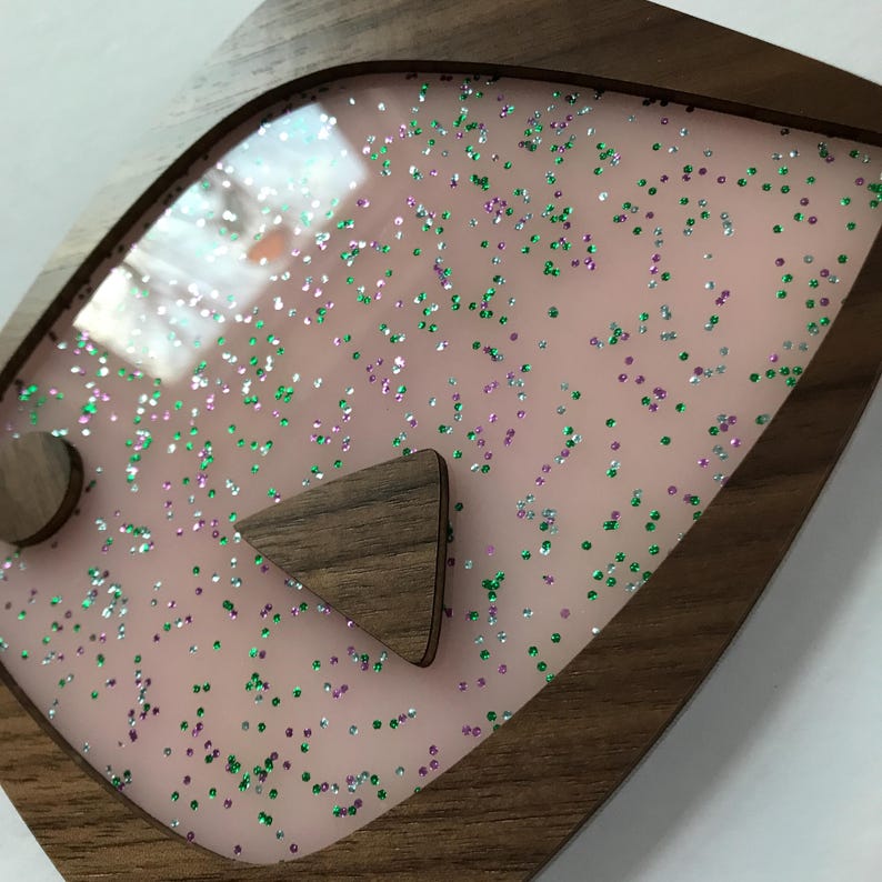 Puede incluir: Un marco decorativo de madera con un interior rosa salpicado de purpurina verde, morada y plateada. Dos formas de madera, un c&iacute;rculo y un tri&aacute;ngulo, est&aacute;n fijadas a la superficie rosa. El marco tiene un acabado de madera oscura.