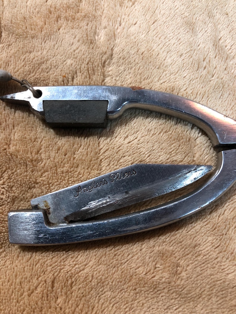 May include: A silver-colored, multi-tool pliers with a small knife blade and a sharpening stone. The tool is engraved with the words "Angler's Pliers".