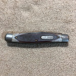 May include: A closed pocket knife with a brown handle and silver accents. The blade is folded into the handle. The text "OLD TIMER" is visible on the handle. The knife is resting on a textured surface.
