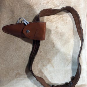 May include: A brown leather belt with a buckle and a brown leather holster attached to it. The holster has a snap closure and the text "WILDERNESS" embossed on it.