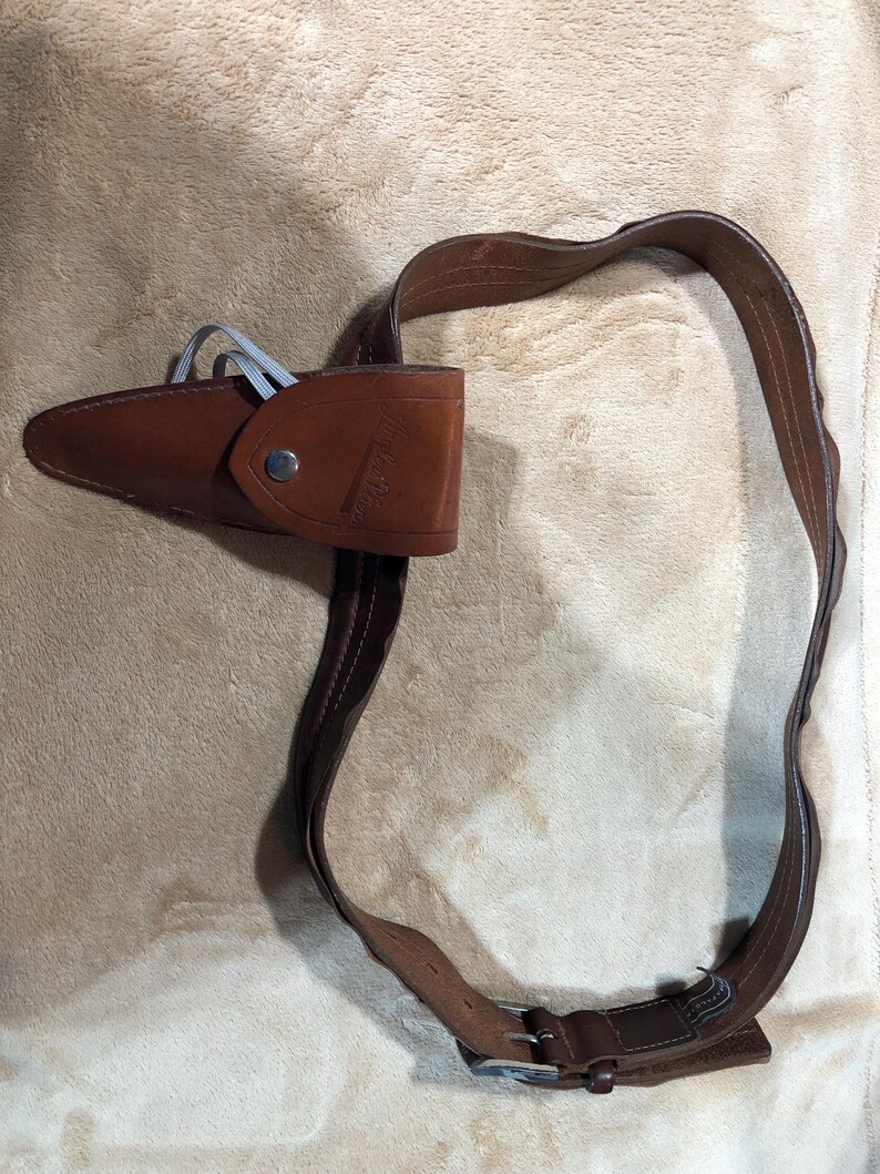 May include: A brown leather belt with a buckle and a brown leather holster attached to it. The holster has a snap closure and the text "WILDERNESS" embossed on it.