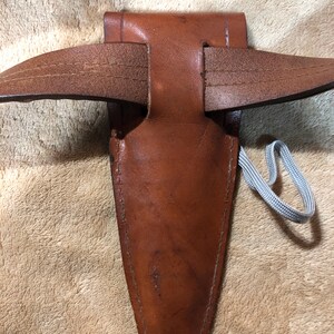 May include: A brown leather sheath with a belt loop and a gray cord. The sheath is designed to hold a knife or other sharp object.
