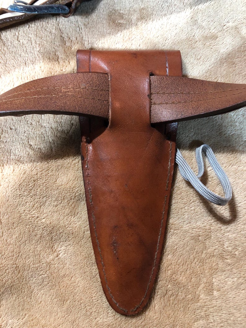 May include: A brown leather sheath with a belt loop and a gray cord. The sheath is designed to hold a knife or other sharp object.