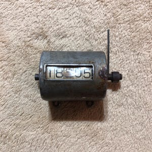 May include: A vintage, cylindrical mechanical counter with a metal casing. The counter displays the number 18505 in white digits. A small lever and a reset knob are visible on the side. The counter shows signs of age and wear.