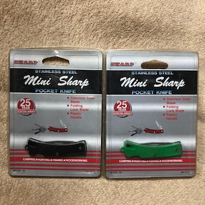 May include: Two Mini Sharp pocket knives in packaging. One has a black handle, the other a green handle. The packaging includes the text "Stainless Steel", "Pocket Knife", and "Camping, Hunting, Fishing, Woodworking".