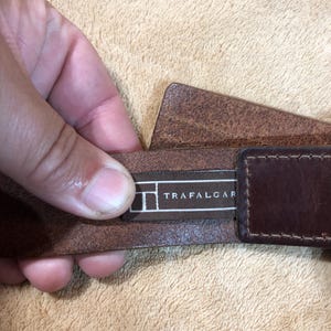 May include: A brown leather belt with a brown leather buckle. The belt has a black label with the text "TRAFALGAR" on it.
