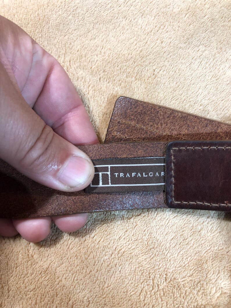May include: A brown leather belt with a brown leather buckle. The belt has a black label with the text "TRAFALGAR" on it.