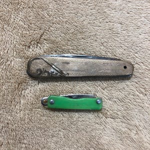 May include: Two pocket knives. The larger knife has a gold-colored metal casing with etched floral designs. The smaller knife has a bright green handle. Both knives are closed, showing their blades tucked inside.
