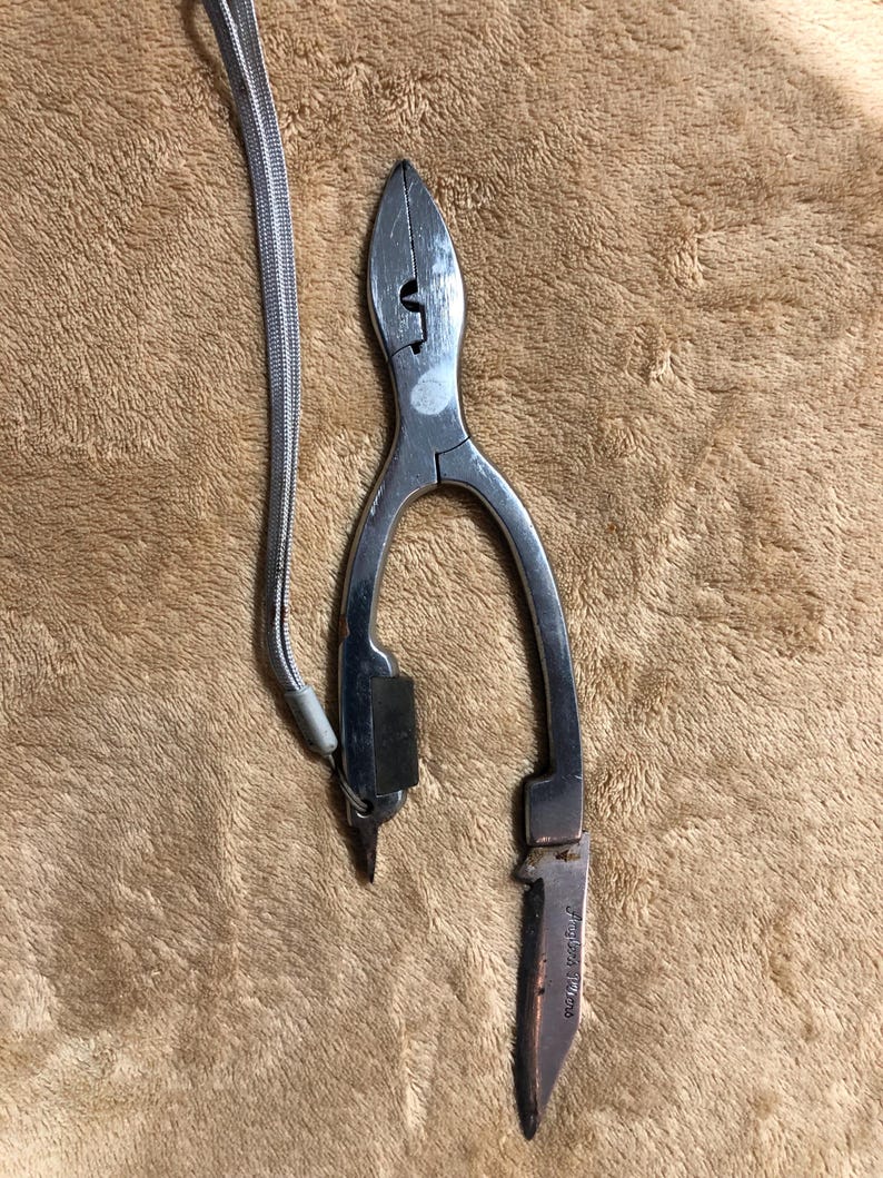 May include: Silver fishing pliers with a small knife attached. The pliers have a lanyard attached to them.