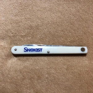 May include: A white pocket knife with the word "Snokist" printed in blue. The knife is closed and has a silver blade. The handle is white with silver rivets and a silver end cap.