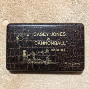 May include: A rectangular brown case with a textured, crocodile-like pattern. The case features the text "CASEY JONES & CANNONBALL" and an illustration of a train. The words "ENGINE 382" and "Frost Cutlery" are also visible.