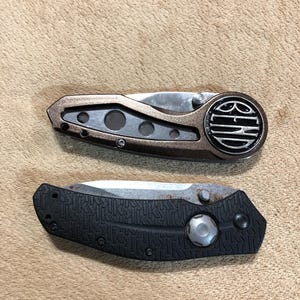 May include: Two folding pocket knives. The top knife has a bronze-colored handle with a circular design on the end. The bottom knife has a black textured handle. Both knives have silver blades.
