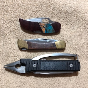 May include: Three folding tools: a top knife with a dark textured handle and turquoise inlay, a middle knife with a wooden handle and brass bolsters, and a bottom multi-tool with black textured grips labeled SPORT MECHANIC TM with pliers.