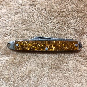 May include: A small, folding pocket knife with a gold glitter handle. The blade is silver, and the handle is secured with three silver rivets. The knife has a small loop at one end for attaching to a keychain.