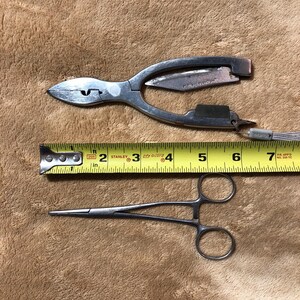 May include: Two surgical tools, a pair of pliers with a small knife and a pair of hemostats, are laid out on a brown fuzzy surface. A yellow measuring tape is placed between the tools. The tape reads 2, 3, 4, 5, 6, and 7 inches.